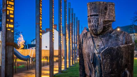 Castle Park in Northampton, with wooden poles with writing on them, a wooden knight and a play area. There is grass on the ground behind the knight.