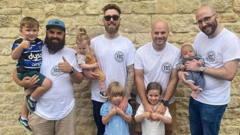 Wayne, Ben, Max and Alex stand together with their children in a group photo. They are all smiling at the camera, and wearing white t-shirts with a black circular FDS logo. 