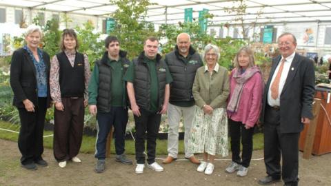 A group photo of two members of Grow standing alongside six members of the Cornish-based Burncoose Nurseries. In the background is the garden.