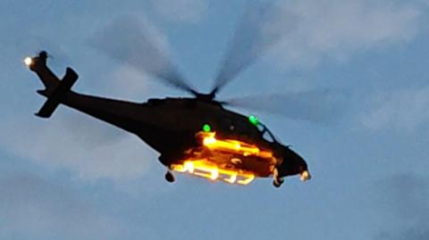 A helicopter in mid-air, seen from the ground and silhoutted against a blue sky with fading light.