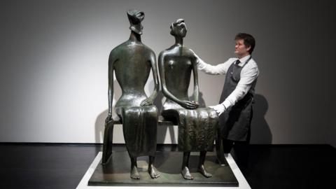 A bronze sculpture of a couple sitting on a bench. A man in white shirt and apron poses with his hands on the sculpture