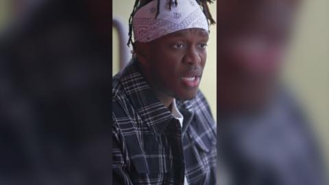 KSI is wearing a white bandana and a blue shirt. He looks off camera.