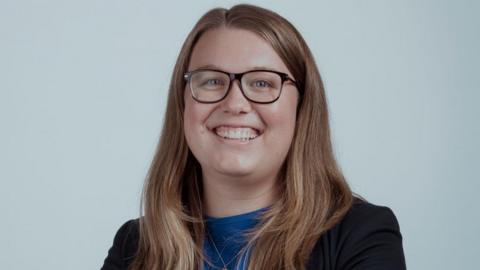 Rachel Hayden is pictured. She has long brown hair and glasses. She is wearing a blue shirt with a black blazer over the top. She is looking at the camera and smiling. 