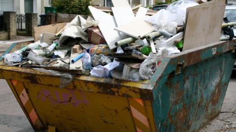 Yellow and blue skip on a street filled with household waste including boarding, pipes and packaging