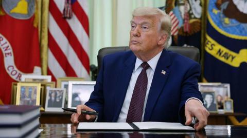 Donald Trump sits in the Oval Office with his hands holding a notepad of paper and a pen. He is wearing a navy suit and burgundy tie. 