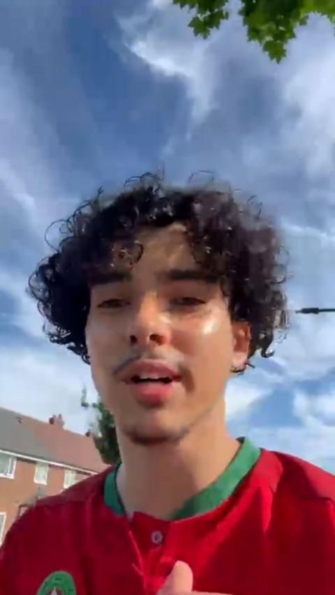 Young man with dark curly hair and red football shirt with blue sky and wispy clouds behind.