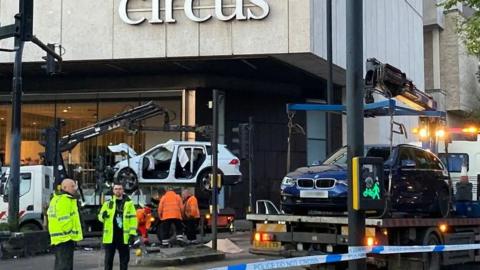 Members of a vehicle recovery team wearing yellow and orange hi-vis jackets. They are standing near the scene of the crash where a white Honda Jazz is being taken away on the bed of a crane truck. The blue BMW is visible on the right, also being removed from the scene.