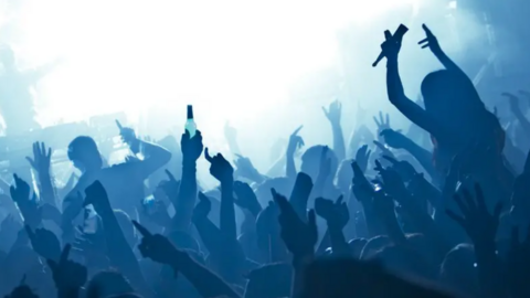 Ravers crowd in front of a DJ on a stage. Some of the dancers are on people's shoulders. Some are holding bottles in the air.  They are silhouetted by the glaring lights. The picture has ben taken from behind the dancers.