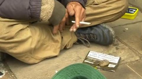 A person sitting crossed-legged on a pavement. They are wearing beige trousers and a boot is visible. They are holding a rolled-up cigarette. On the ground in front of them are a few coins on a cigarette packet and upturned, green hat. 