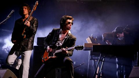 Three members of Arctic Monkeys on a smoky stage at Glastonbury at night. On the left, a man plays a bass guitar, the neck raised towards the sky. He has bushy brown hair and wears a black leather jacket and light-grey trousers. In the centre, a man with brown hair plays a brown guitar while looking to his left. He wears sunglasses, a dark suit and white shirt, unbuttoned at the chest. To the right, a man with short hair bends over a keyboard, his right hand on the keys. 