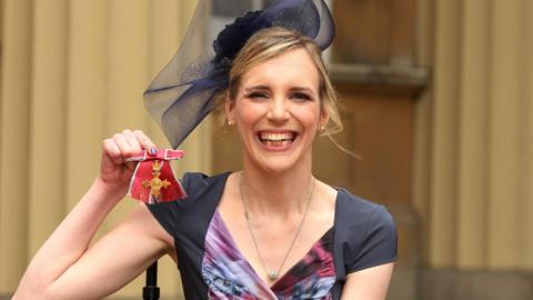 Sophie Christiansen smiling at the camera with a navy fascinator on her head, wearing a navy and purple dress and holding her medal up  in her left hand.