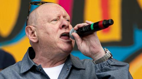 Shaun Ryder, with sunglasses perched on his bald head, sings into a microphone while wearing a large silver watch, denim shirt and white t-shirt. The blurred background is part of a yellow, red, black and blue graphic of the words Happy Mondays