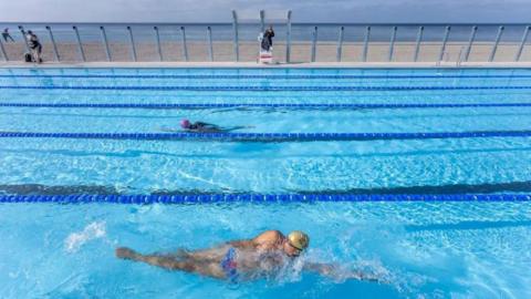 People swimming in a swimming pool with a lifeguard on duty. In the background is Brighton beach.