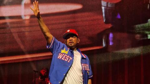 Will Smith in a white T-shirt and open blue baseball shirt with West Phil on it, and wearing a red baseball cap slightly twisted to the right. He is on stage and has his right hand raised in the air. He looks a little out of breath and sweaty. The stage behind him is backlit in red.