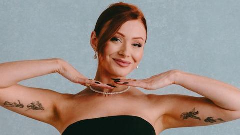 Elz the Witch poses in front of a light blue background. She's wearing a strapless black gown and a thing string of pearls. She's smiling, her hands under her chin, framing her face, and showing off the tattoos of figures drawn in intricate black ink lines on the inside of her upper arms.