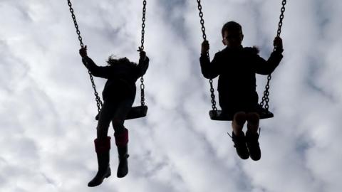 The shadowed outline of two children swinging high on swings with clouds in the background