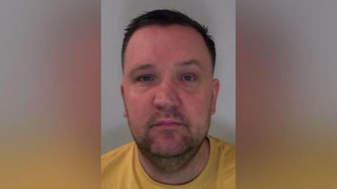 Mugshot of a man in a yellow t-shirt with stubble and short dark hair