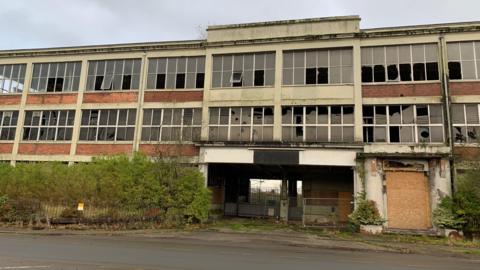 A dilapidated factory building with smashed windows, plants growing out of the roof and a boarded-up entrance way