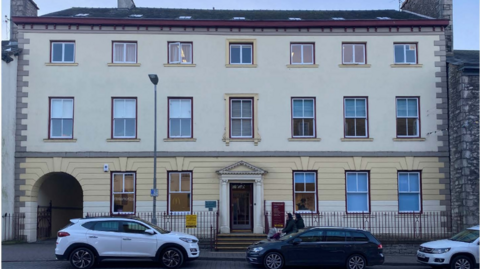 Strickland House is a Georgian building which has large windows and decorative panels. It is painted cream, white and maroon and looks well-cared for. There are two cars parked outside.
