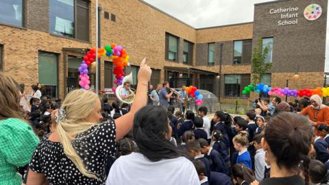 School children dance to a brass band along with teachers, colourful balloons decorate the front of the new school 