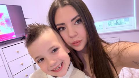 a boy with a smart waistcoast and bow tie smiles as he is hugged by his mum who has long brown hair and is pouting