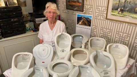 An elderly woman with short grey hair sits behind a kitchen table which has about 10 bedpans resting on it in all different shapes and sizes. The woman is wearing a white blouse and has glasses. She is smiling at the camera.