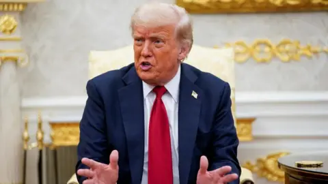 Donald Trump sitting in a gold chair in a room with gold decoration, wearing a red tie and a blue jacket and holding his hands out in front of him
