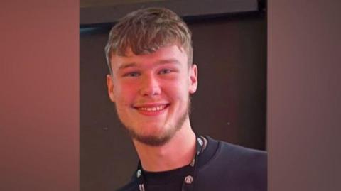 A young man with short light brown hair and a beard is smiling. He is wearing a navy coloured top and a lanyard. 