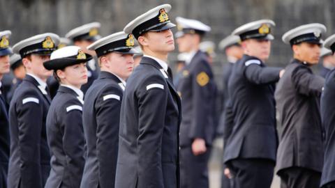 A cohort of Royal Navy recruits stands on parade during a passing out ceremony. They are all wearing uniform with hats on.