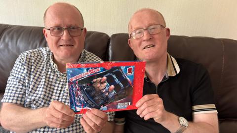 Two older adults with short or thinning hair and glasses sit on a brown leather sofa, wearing casual short‑sleeve shirts, holding a souvenir photo card from Pleasure Beach showing a roller coaster ride with the text “We Survived the Big One” and “Big 1,” against a light textured wall.
