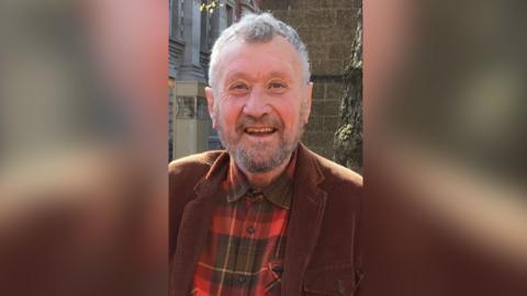 A man with grey hair and a grey beard smiling at the camera. He is wearing a brown jacket and a red and black checked shirt