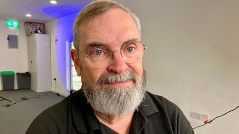 A man with a grey beard wearing glasses and a black polo shirt.