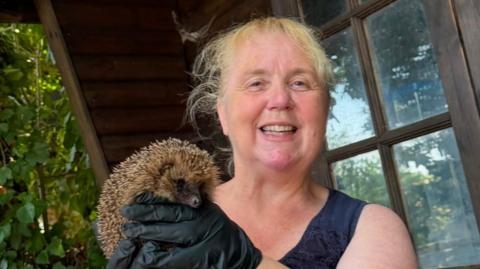 Paula Baker holding a hedgehog while smiling. She has black latex gloves on and a navy sleeveless top.