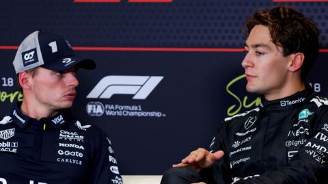 George Russell speaking to Max Verstappen at an FIA-F1 news conference during the 2025 Singapore Grand Prix
