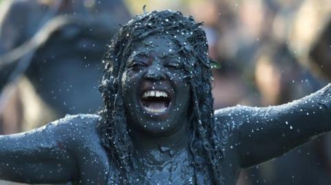 A woman laughing whilst covered completely in mud, holding her arms out to the sides