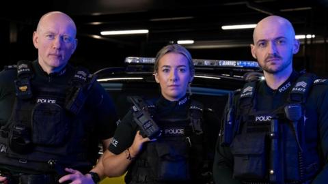 Three police officers looking at the camera. All are wearing police uniform that is black and dark green. The one on the left is a bald man, in the middle is a blonde woman and on the right is a bald man with dark beard.