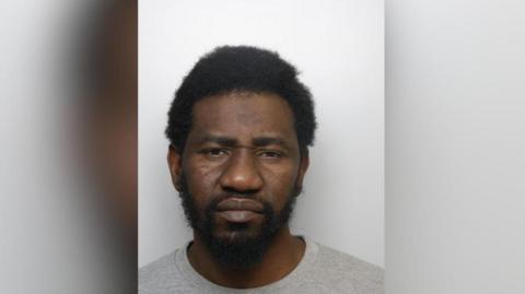 A mugshot of Robert Jean. A man with black Afro-hair and stubble staring into the camera. He is wearing a grey shirt.