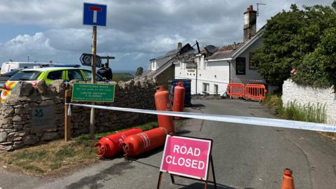 The entrance to the Worm's Head Hotel. There is a road closed sign just at the entrance with police table above it. There is orange canister lying on the ground. The roof of the hotel has been damaged by the fire. There is a police car parked in front of the hotel