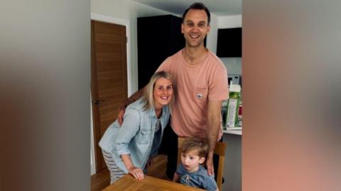 A woman, man and a young boy. The young boy is wearing a blue t-shirt and is sitting at a table. The woman is leaning into the photo and is wearing a denim top. The man standing is wearing a pink top. The photo has been taken at a dining table with the kitchen in the background.