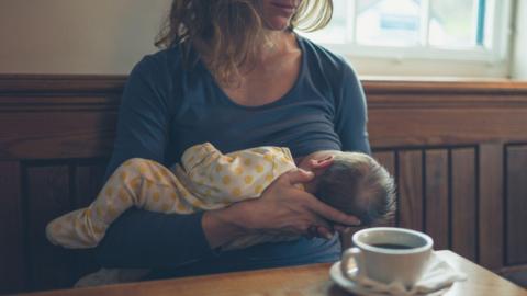 In a cafe with dark wooden backed benches, a woman in a blue top breastfeeds a baby in a yellow and white babygrow. A cup of coffee in a white cup sits on a table in front of them.