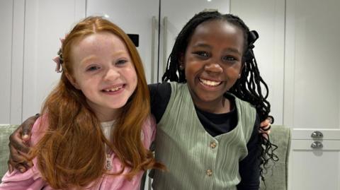 Two girls are hugging whilst smiling at the camera. One girl is wearing pink and the other girl is wearing green. 