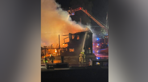 Firefighters at the scene of a fire at an industrial building in Standard Quay, Faversham, Kent. Patches of fire can be seen in the building, alongside a crane with a hose spraying the flames. Several firefighters can be seen in the image.