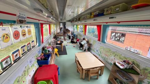 A converted railway carriage which has been turned into classroom space for a pre-school. There are tables and chairs on the floor, colourful drawings along the wall and other teaching materials.