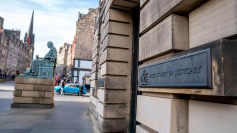 A general view of the outside of the High Court in Edinburgh. It is a stone building and the view is looking up the street to a statue and other buildings