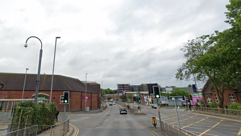 A dual carriageway, with buildings lining both sides. Traffic lights in the foreground are on green, with several cars in the distance. The carriageways are separated by railings on a  concrete section. There is a part-built building in the distance several storeys high