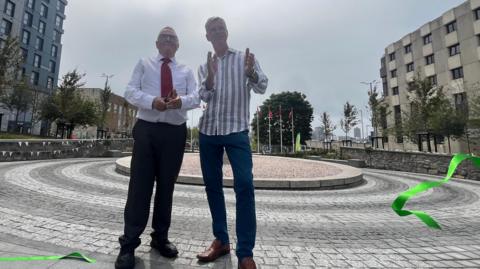 Two men stand smiling and dressed smartly in a round stone-paved area. One holds a pair of scissors and there is a green ribbon floating to ground as it has just been cut. The other man is applauding. 