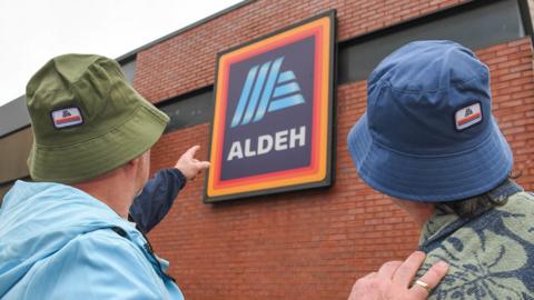 Two men wearing bucket hats, one green and one blue, stand with their back to the camera and point at the Aldi sign, which features the shop's orange and red bordered blue logo and the word Aldeh, on the side of one their brick built store