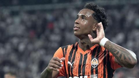 Kevin puts his hand to  his ear in an orange and black striped Shakhtar shirt