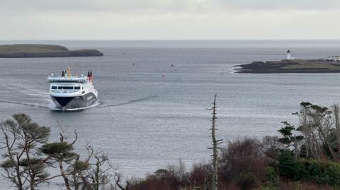 A white boat with a black hull sails on blue/grey waters, with two pieces of green and brown land either side, including a white lighthouse on the rigt. Trees and bushes fill the bottom half of the picture, with grey skies in the distance. 