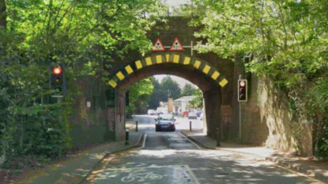 A car drives towards the camera on as the road narrows to one lane under a brick railway bridge. There are traffic lights showing red in the foreground and greenery around the bridge